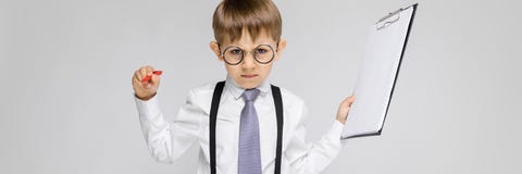 Очаровательный мальчик в белой рубашке, подтяжках, связи и джинсах света стоит на серой предпосылке Мальчик держит ручку и стоковая фотография Очаровательный мальчик в белой рубашке, подтяжках, связи и джинсах света стоит на серой предпосылке Мальчик держит ручку и стоковая фотография