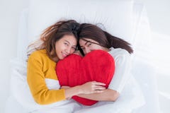 Положение пар Азии лесбосское LGBT на радуге кровати и объятия красит форму сердца подушки с моментом счастья совместно, концепци стоковые фотографии rf Положение пар Азии лесбосское LGBT на радуге кровати и объятия красит форму сердца подушки с моментом счастья совместно, концепци стоковые фотографии rf