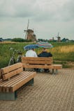 Поля и ветрянка пожилых пар сидя наблюдая на Kinderdijk стоковое фото rf Поля и ветрянка пожилых пар сидя наблюдая на Kinderdijk стоковое фото rf