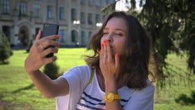 Портрет девушки детенышей усмехаясь кавказской стоя в парке и используя smartphone, делая selfie, университет в акции видеоматериалы Портрет девушки детенышей усмехаясь кавказской стоя в парке и используя smartphone, делая selfie, университет в акции видеоматериалы