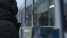 Портрет конца-вверх молодого длинн-с волосами человека с бородой в наушниках стоя на трамвайной остановке в зиме и ожидании сток-видео Портрет конца-вверх молодого длинн-с волосами человека с бородой в наушниках стоя на трамвайной остановке в зиме и ожидании сток-видео
