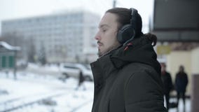 Портрет молодого длинн-с волосами человека с бородой в наушниках стоя на трамвайной остановке в зиме и ждать трамвай сток-видео Портрет молодого длинн-с волосами человека с бородой в наушниках стоя на трамвайной остановке в зиме и ждать трамвай сток-видео