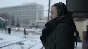 Портрет молодого длинн-с волосами человека с бородой в наушниках стоя на трамвайной остановке в зиме и ждать трамвай сток-видео Портрет молодого длинн-с волосами человека с бородой в наушниках стоя на трамвайной остановке в зиме и ждать трамвай сток-видео