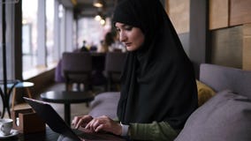 Портрет усмехаясь молодой мусульманской женщины работая на современном ноутбуке в кафе Привлекательная женщина в hijab раскрывает видеоматериал Портрет усмехаясь молодой мусульманской женщины работая на современном ноутбуке в кафе Привлекательная женщина в hijab раскрывает видеоматериал