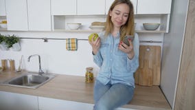 Привлекательная девушка использует умный телефон и ест яблоко, сидя на kitc стоковое фото Привлекательная девушка использует умный телефон и ест яблоко, сидя на kitc стоковое фото
