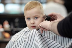 Серьезный и маленький вспугнутый милый белокурый ребенок с голубыми глазами в парикмахерской имея моя голову парикмахером стоковое изображение rf Серьезный и маленький вспугнутый милый белокурый ребенок с голубыми глазами в парикмахерской имея моя голову парикмахером стоковое изображение rf