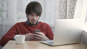Счастливый фрилансер молодого человека работая с современными компьтер-книжкой и телефоном в кафе принимая чашку кофе сток-видео Счастливый фрилансер молодого человека работая с современными компьтер-книжкой и телефоном в кафе принимая чашку кофе сток-видео