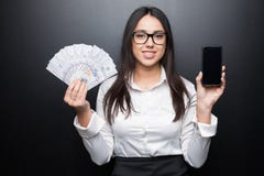 Счастливая молодая женщина брюнет в белой рубашке показывая smartphone с пустым экраном и деньгами наличных денег в руках на черн стоковые фотографии rf Счастливая молодая женщина брюнет в белой рубашке показывая smartphone с пустым экраном и деньгами наличных денег в руках на черн стоковые фотографии rf