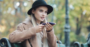 Счастливая молодая женщина используя функцию опознавания голоса смартфона Она сидя на стенде в парке и предписаниях осени стоковое изображение Счастливая молодая женщина используя функцию опознавания голоса смартфона Она сидя на стенде в парке и предписаниях осени стоковое изображение