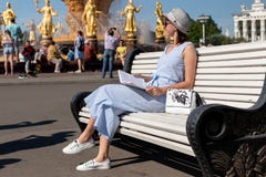 Счастливая молодая и стильная женщина со шляпой и книгой сидя на стенде в парке стоковое изображение rf Счастливая молодая и стильная женщина со шляпой и книгой сидя на стенде в парке стоковое изображение rf
