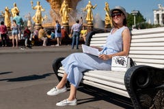 Счастливая молодая и стильная женщина со шляпой и книгой сидя на стенде в парке стоковая фотография rf Счастливая молодая и стильная женщина со шляпой и книгой сидя на стенде в парке стоковая фотография rf