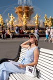 Счастливая молодая и стильная женщина со шляпой и книгой сидя на стенде в парке стоковая фотография rf Счастливая молодая и стильная женщина со шляпой и книгой сидя на стенде в парке стоковая фотография rf