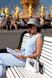 Счастливая молодая и стильная женщина со шляпой и книгой сидя на стенде в парке стоковые фото Счастливая молодая и стильная женщина со шляпой и книгой сидя на стенде в парке стоковые фото