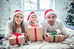 Счастливые мать, отец и маленькая девочка в шляпах хелпера santa с подарочными коробками над предпосылкой живущей комнаты и рожде стоковые изображения Счастливые мать, отец и маленькая девочка в шляпах хелпера santa с подарочными коробками над предпосылкой живущей комнаты и рожде стоковые изображения