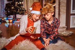 Счастливый брат и сестра нося Санта и шляпы оленей сидя на ковре меха и раскрывают подарок около рождественской елки на стоковая фотография Счастливый брат и сестра нося Санта и шляпы оленей сидя на ковре меха и раскрывают подарок около рождественской елки на стоковая фотография