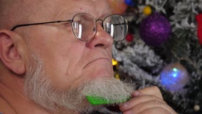 Счастливый пожилой человек расчесывая его серую бороду с гребнем на предпосылке рождественской елки в гирляндах, зеленых шариках сток-видео Счастливый пожилой человек расчесывая его серую бороду с гребнем на предпосылке рождественской елки в гирляндах, зеленых шариках сток-видео