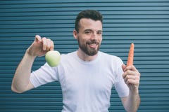 Счастливый человек стоящ и держащ фрукт и овощ в руках Это зеленая морковь яблока и апельсина Он смотрит дальше стоковые фотографии rf Счастливый человек стоящ и держащ фрукт и овощ в руках Это зеленая морковь яблока и апельсина Он смотрит дальше стоковые фотографии rf
