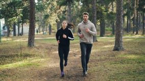 Тележка снятая привлекательной девушки и парня jogging в парке совместно сфокусированном на практике Женщина носит фуфайку и видеоматериал Тележка снятая привлекательной девушки и парня jogging в парке совместно сфокусированном на практике Женщина носит фуфайку и видеоматериал