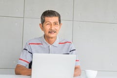 Усмехающся, азиатский старший человек Платье со случайными одеждами сидя и работая на таблице с кружкой ноутбука и белого кофе, стоковые изображения rf Усмехающся, азиатский старший человек Платье со случайными одеждами сидя и работая на таблице с кружкой ноутбука и белого кофе, стоковые изображения rf