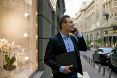 Успешный бизнесмен в костюме с компьтер-книжкой говоря на телефоне и усмехаясь в городе стоковое изображение Успешный бизнесмен в костюме с компьтер-книжкой говоря на телефоне и усмехаясь в городе стоковое изображение