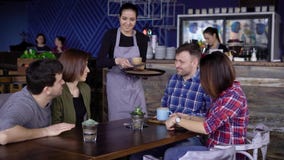 Хлебосольная молодая официантка на кафе принося напитки в покрашенных кружках к клиентам 4 друз сидя на акции видеоматериалы Хлебосольная молодая официантка на кафе принося напитки в покрашенных кружках к клиентам 4 друз сидя на акции видеоматериалы