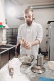 Харизматический хлебопек с бородой и усиком замешивает тесто на таблице в пекарне стоковая фотография rf Харизматический хлебопек с бородой и усиком замешивает тесто на таблице в пекарне стоковая фотография rf