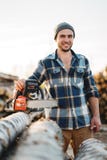 Цепная пила владением рубашки шотландки сильного бородатого woodcutter нося в руке на предпосылке лесопилки и складе деревьев стоковые фотографии rf Цепная пила владением рубашки шотландки сильного бородатого woodcutter нося в руке на предпосылке лесопилки и складе деревьев стоковые фотографии rf