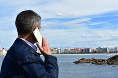 Человек говоря на телефоне Одежды голубых джинсов, белый смартфон Прибрежная деревня с пляжем, утесами и прогулкой стоковые изображения Человек говоря на телефоне Одежды голубых джинсов, белый смартфон Прибрежная деревня с пляжем, утесами и прогулкой стоковые изображения