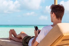 Человек на sunchair в тропическом положении используя его смартфон Ясная вода бирюзы как предпосылка стоковые фото Человек на sunchair в тропическом положении используя его смартфон Ясная вода бирюзы как предпосылка стоковые фото