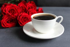 Черный кофе в белой чашке с поддонником на таблице, букете красных роз стоковые изображения Черный кофе в белой чашке с поддонником на таблице, букете красных роз стоковые изображения