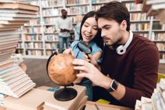 Этническая азиатская девушка и белый парень окруженные книгами в библиотеке Студенты используют глобус стоковое изображение rf Этническая азиатская девушка и белый парень окруженные книгами в библиотеке Студенты используют глобус стоковое изображение rf