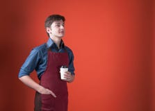 与黑发的英俊的barista在拿着纸杯用咖啡和看在珊瑚颜色背景的红色围裙 免版税图库摄影 与黑发的英俊的barista在拿着纸杯用咖啡和看在珊瑚颜色背景的红色围裙 免版税图库摄影