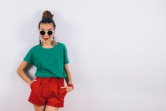 佩带在红色短裤和绿色T恤杉的太阳镜的美丽的行家妇女站立在白色墙壁和微笑附近 免版税库存图片 佩带在红色短裤和绿色T恤杉的太阳镜的美丽的行家妇女站立在白色墙壁和微笑附近 免版税库存图片