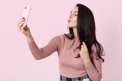 使用智能手机的迷人的女孩照片为selfie 有长的黑发的可爱的女性做selfie,保持嘴唇被环绕,妇女 免版税库存图片 使用智能手机的迷人的女孩照片为selfie 有长的黑发的可爱的女性做selfie,保持嘴唇被环绕,妇女 免版税库存图片