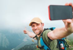拍selfie照片使用智能手机和显示赞许的年轻背包徒步旅行者人在走由有雾的多云天气期间 免版税库存图片 拍selfie照片使用智能手机和显示赞许的年轻背包徒步旅行者人在走由有雾的多云天气期间 免版税库存图片