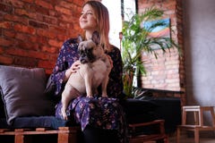 礼服的愉快的白肤金发的妇女与她逗人喜爱的哈巴狗坐一个手工制造沙发在有顶楼内部的屋子里 库存图片 礼服的愉快的白肤金发的妇女与她逗人喜爱的哈巴狗坐一个手工制造沙发在有顶楼内部的屋子里 库存图片