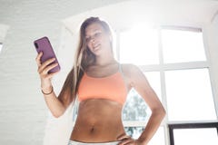 被晒黑的运动的妇女佩带的sportwear和无线耳机在白色晴朗的健身房拿着智能手机手中并且计数卡路里在fi 免版税图库摄影 被晒黑的运动的妇女佩带的sportwear和无线耳机在白色晴朗的健身房拿着智能手机手中并且计数卡路里在fi 免版税图库摄影