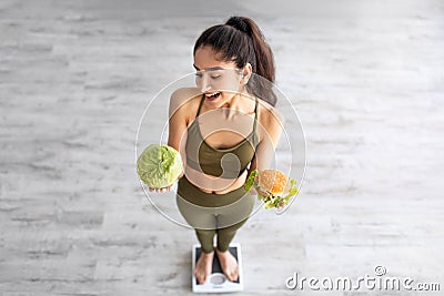 Above view of cheerful Indian lady holding cabbage and hamburger on scales indoors, full length Stock Photo