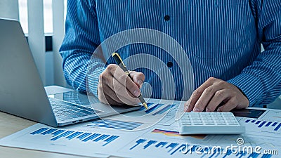 An accountant using a calculator and computer with a pen in hand is making money and analyzing graph papers on the table about exp Editorial Stock Photo