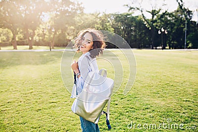 Active brunette girl with short hair is walking in park on sunset. She wears white T-shirt, shirt and jeans, holds bag Stock Photo