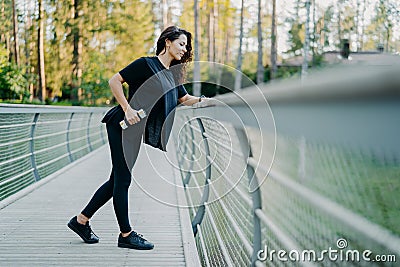 Active brunette woman dressed in casual t shirt and leggings, holds dumbbells, poses at bridge, looks down, goes in for sport Stock Photo