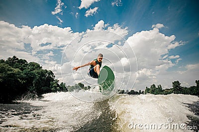 Active man wakesurfing on the board down the river against the cloudy sky and trees Stock Photo