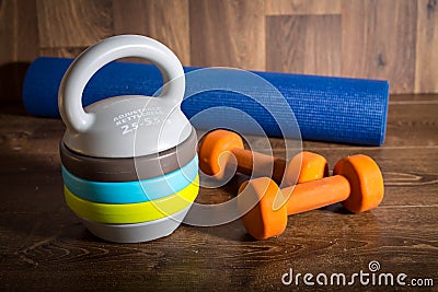 Adjustable kettlebell, pair of orange dumbbells and yoga mat on wooden background. Weights for a fitness training. Stock Photo