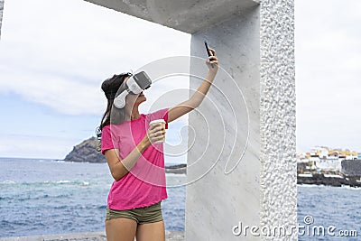 Adolescent taking selfie with virtual reality glasses with her cellphone while taking a take away coffee in the sea, to sharing in Stock Photo