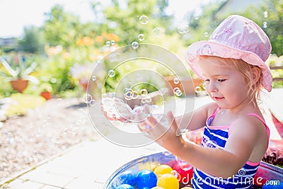 Adorable blonde baby girl 3 year old in a pink hat and blue stripped swimsuit having bath at backyard and playing with bubbles Stock Photo