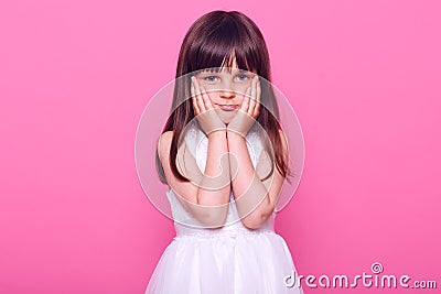 Adorable bored small girl looking at camera with tired expression, keeping hands on cheeks, does not know what to do, wearing Stock Photo