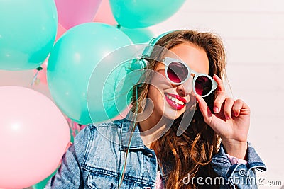 Adorable brunette young woman looking through stylish sunglasses and posing with smile next to colorful balloons. Close Stock Photo