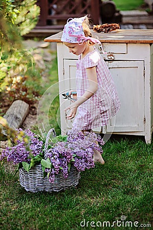 Adorable child girl in pink plaid dress near vintage bureau with lilacs in basket Stock Photo