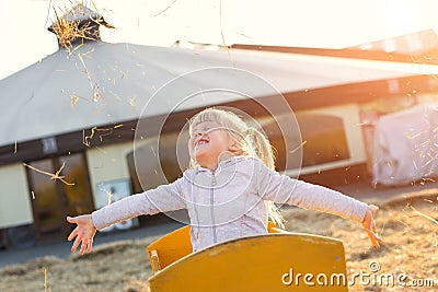 Adorable cute caucasian blond kid girl sitting in wooden cart having fun throwing straw or hay at farm or park during warm autumn Stock Photo