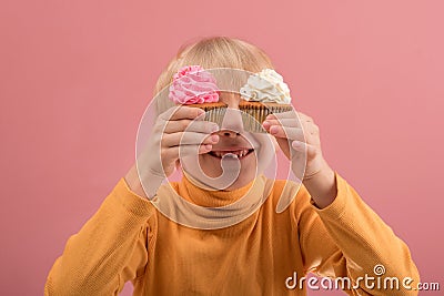 Adorable happy fair-haired boy smiling and holding yummy cupcakes in hands on pink background Stock Photo